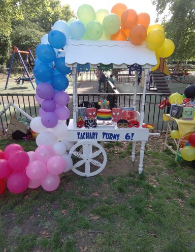 Colourful balloon arch over a cart displaying birthday cakes and treats in a park setting.