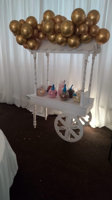 A white cart with colourful drinks, topped with a cluster of gold balloons.