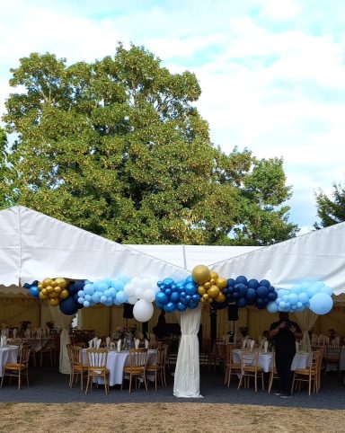 A marquee decorated with blue and gold balloons, set for a dining event.