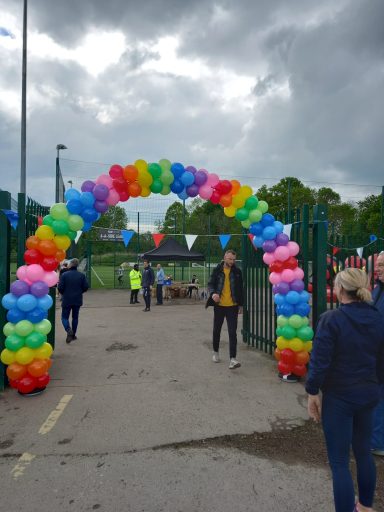 Colourful balloon arch at an event entrance with people nearby and cloudy skies.
